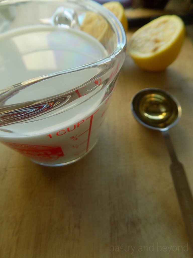 Milk in a measuring glass, lemon cut in half and vinegar in a measuring spoon.