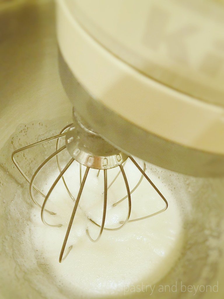 Whisking egg whites and sugar with a whisk attachment in a stand mixer.