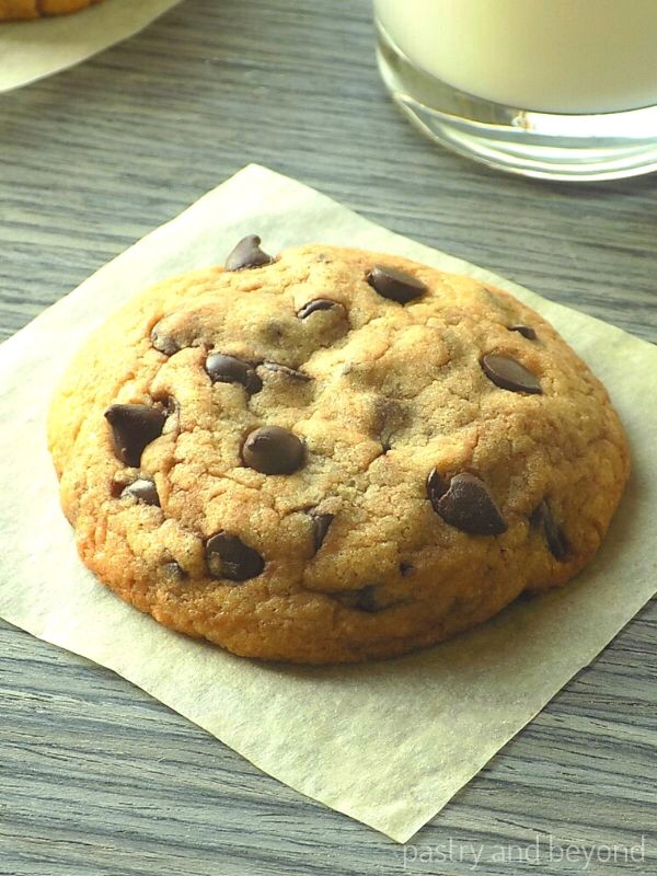 Chocolate chip cookie on a parchment paper with a glass of milk in the background.