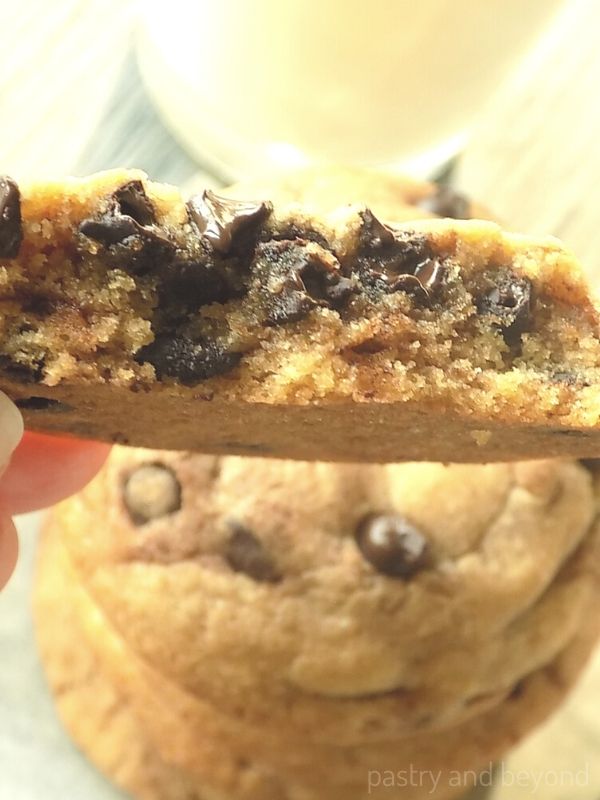 A hand holding half of a soft chocolate chip cookie above the stacked cookies.