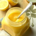 Homemade lemon curd in a jar with a spoon, half lemons in the background.