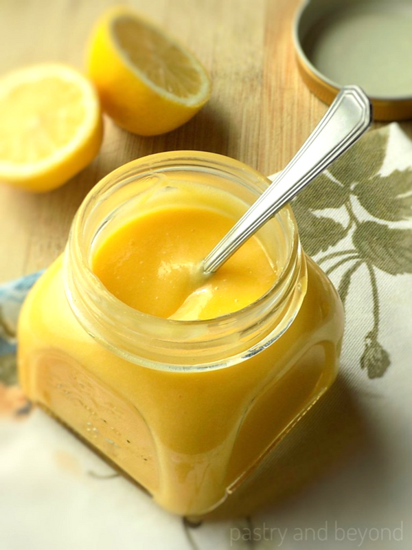 Homemade lemon curd in a jar with a spoon, half lemons in the background.