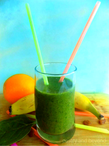 Green iron rich smoothie in a glass with straws, banana and orange in the background.