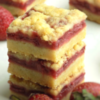 Stacked strawberry crumble bars on a white surface, strawberries in the back and front.