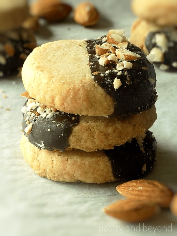 Stacked chocolate dipped almond shortbread cookies with crushed almonds on top on parchment paper.
