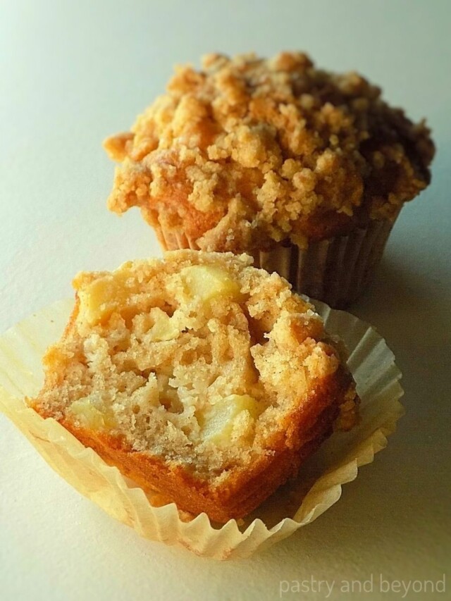 Apple crumble muffin that is cut in half and whole apple muffin in the background.