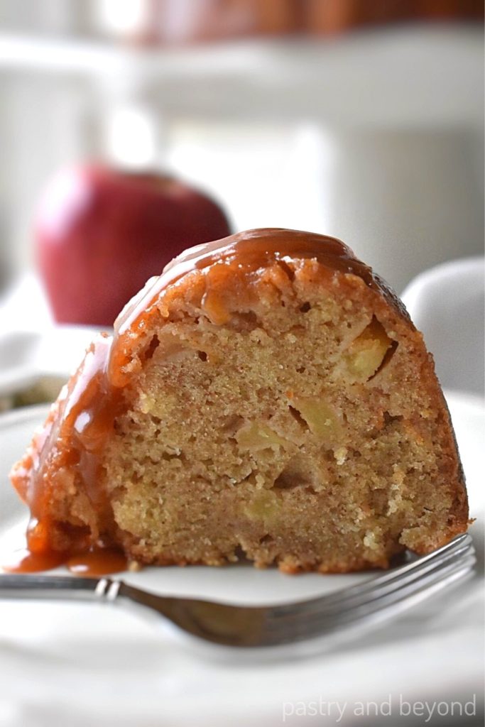A slice of apple bundt cake on a white plate, apples on the background.