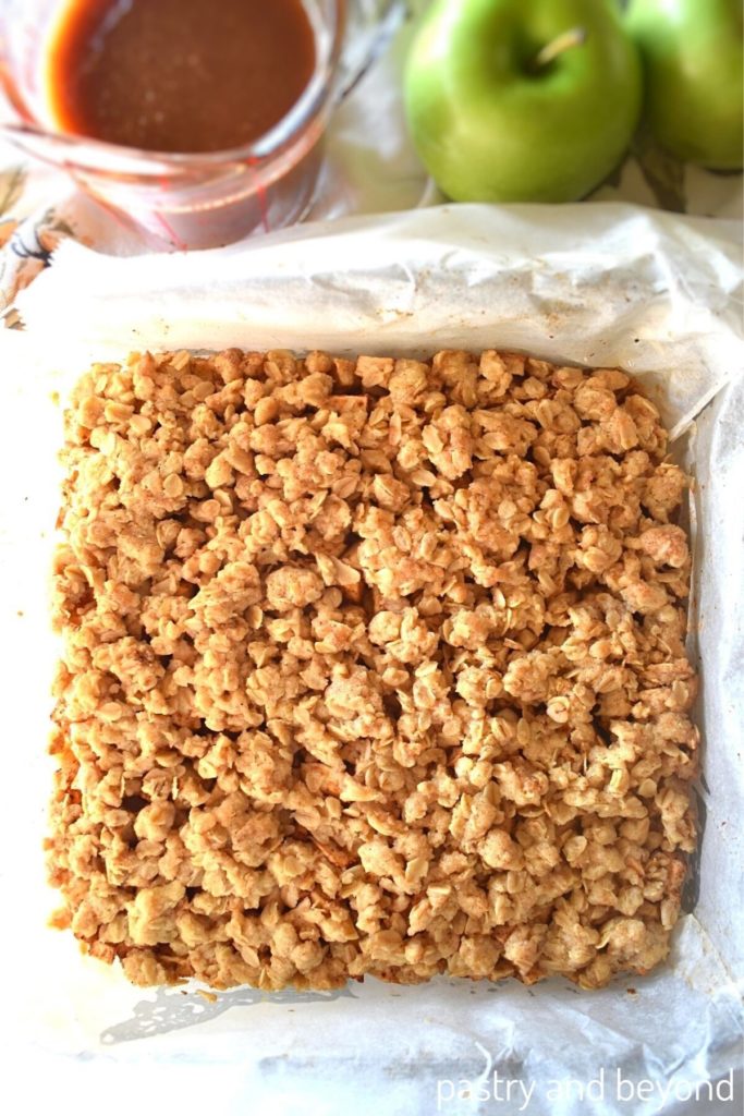 Apple oatmeal bars in a baking dish with caramel sauce and apples in the background.