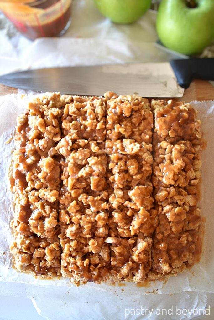 Apple bars that are cut into 16 slices with knife, caramel and apples in the background.