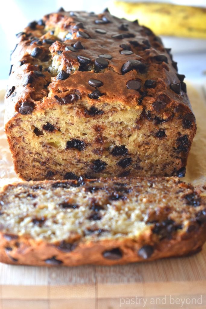 One bowl chocolate chip banana bread loaf on a wooden board with banana in the background. 