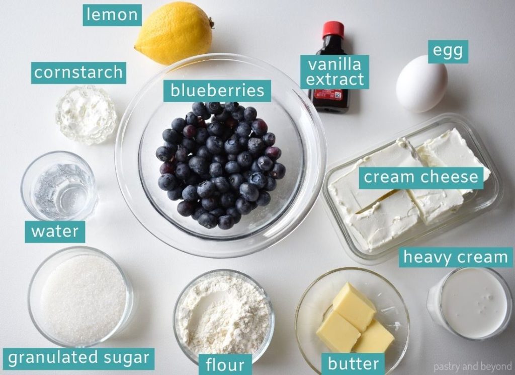 Ingredients to make mini blueberry cheesecakes on a white surface.