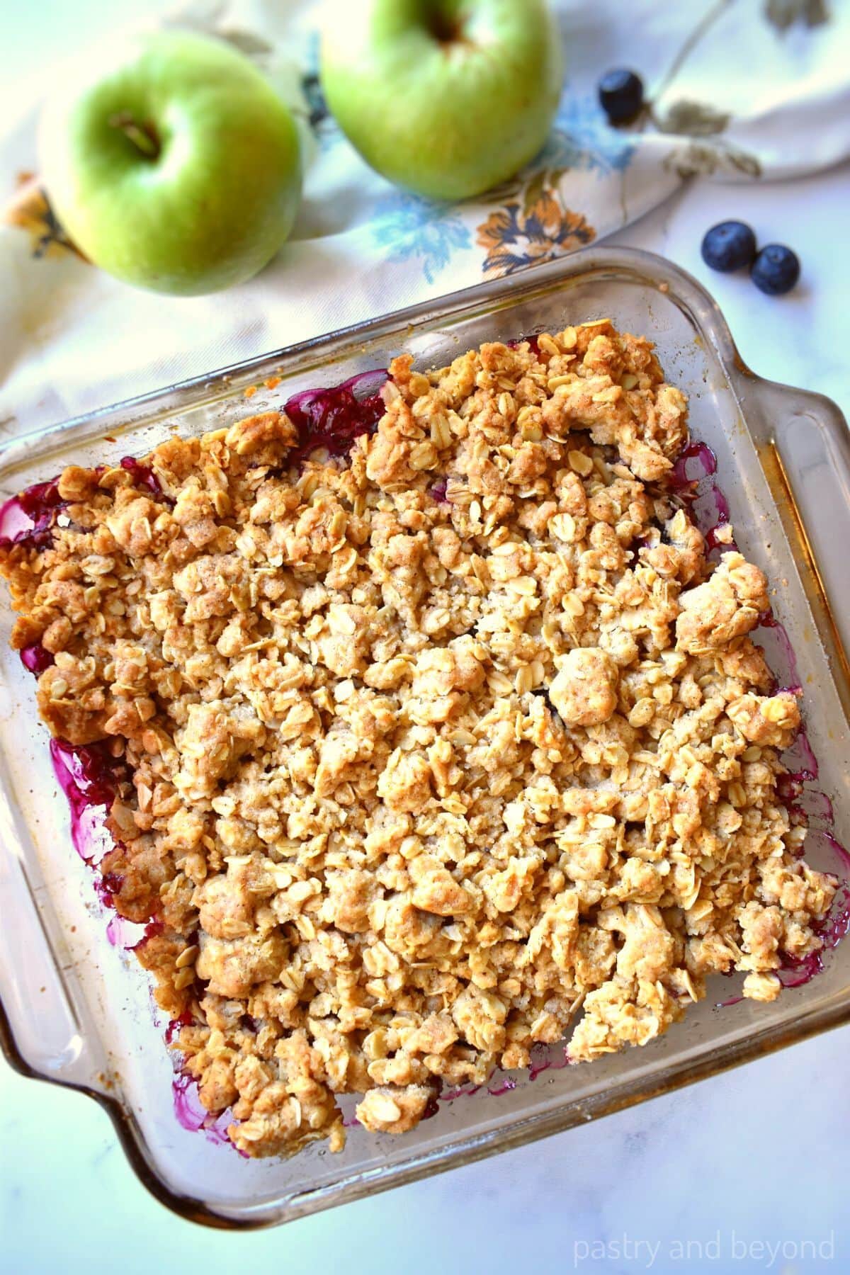 Apple and blueberry crumble in a baking dish with apples and blueberries in the background.