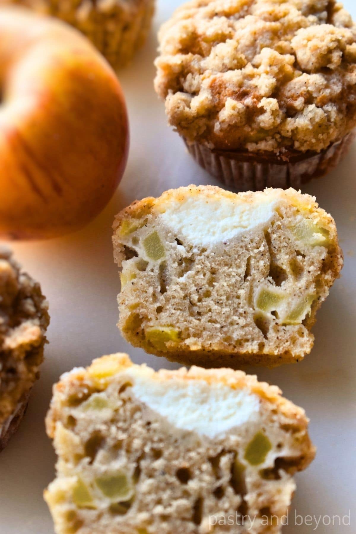 Apple cream cheese muffin that is cut in half and whole muffins on a white surface.